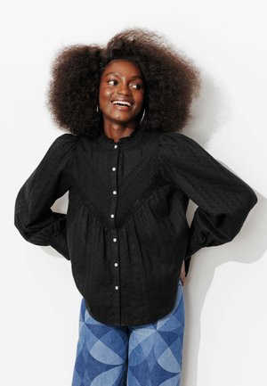 Femme aux cheveux bouclés et volumineux souriante, portant un chemisier noir boutonné à manches bouffantes et un pantalon bleu à motifs, debout devant un fond blanc.