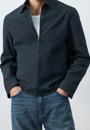 Mand iført en mørk lynlåstrøje over en hvid skjorte med hænderne i lommerne på blå jeans, stående mod en ensfarvet baggrund.