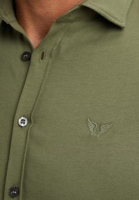 Olijfgroene shirt met korte mouwen van katoen, voorzien van een knoopsluiting aan de voorkant, zwarte knopen en een geborduurd adelaarlogo op de borst.