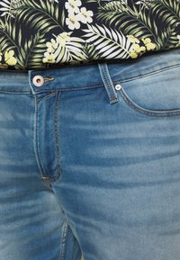 Jeans de mezclilla azul claro con una textura suave, que cuentan con una cremallera, passadores para cinturón y costuras en contraste. Llevados con una camisa de estampado floral.
