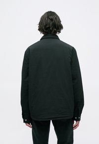 Casaco preto com um corte solto, colarinho clássico e punhos com botões. Feito de um tecido leve com uma textura suave, sem padrões visíveis.