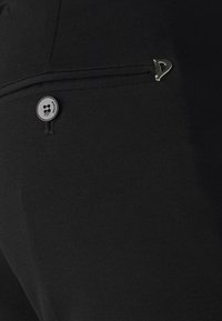 Pantalon en tissu noir avec un design élégant, doté d'une fermeture à bouton et d'un détail métallique près de la poche. Texture lisse.