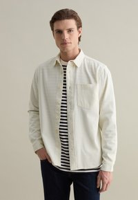 Lichtgekleurde button-up shirt met een zak, gedragen over een zwart-wit gestreept shirt. Donkere jeans maken de outfit compleet. Gladde stoftextuur.