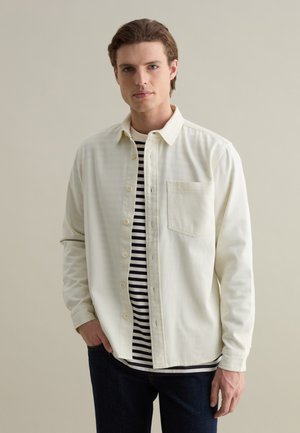 Light-colored button-up shirt with a pocket, worn over a black and white striped shirt. Dark jeans complete the outfit. Smooth fabric texture.