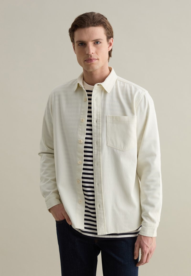 Lichtgekleurde button-up shirt met een zak, gedragen over een zwart-wit gestreept shirt. Donkere jeans maken de outfit compleet. Gladde stoftextuur.