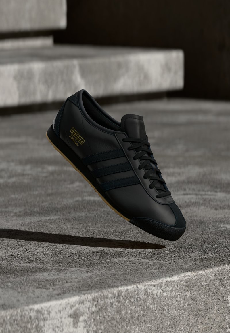 Black leather sneaker with three darker stripes, round toe, and gum rubber sole. Features laces and gold logo detail on the side.