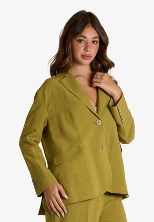 Donna che indossa un blazer oversize verde oliva e pantaloni abbinati, con capelli mossi e sciolti, posa davanti a uno sfondo bianco.