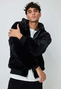 Veste en fausse fourrure noire avec fermeture éclair, capuche, poignets côtelés et coupe décontractée. Portée sur une chemise blanche, mettant en valeur la texture moelleuse.