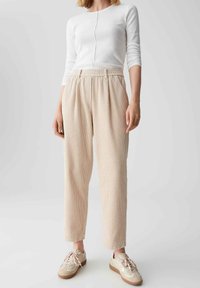 Beige Ripp-Hosen mit elastischem Bund; kombiniert mit einem weißen Langarmoberteil und beigen Sneakers mit hellrosa Schnürsenkeln und Akzenten.