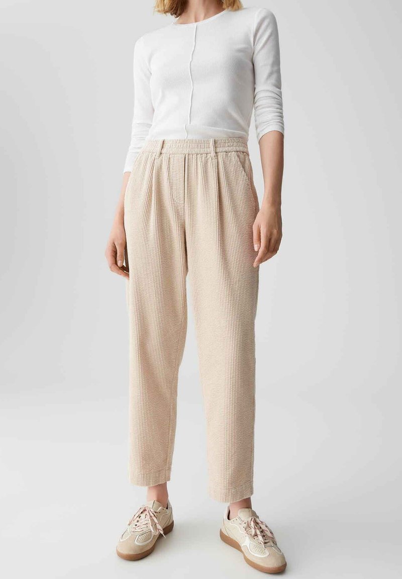 Beige Ripp-Hosen mit elastischem Bund; kombiniert mit einem weißen Langarmoberteil und beigen Sneakers mit hellrosa Schnürsenkeln und Akzenten.