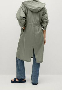 Lichtgroene, waterdichte lange jas met capuchon, getailleerde taille en zijzakken. Gecombineerd met wijd uitlopende blauwe jeans en marineblauwe sandalen.