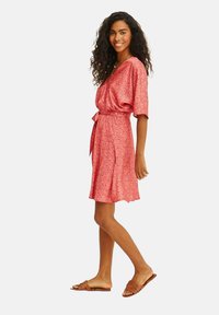 Růžové šaty s krátkým rukávem a délkou do kolen, s malým květinovým vzorem, s přepásaným pasem a detaily na zavázání. V kombinaci s hnědými plochými sandály.