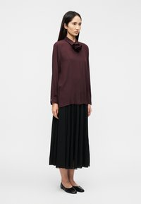 Μπλούζα βουργουνδίας με λουλουδάτο στοιχείο, σε συνδυασμό με μακριά μαύρη πλισέ φούστα και μαύρα μπαλαρίνια, με κλασικό γιακά και μακριά μανίκια.