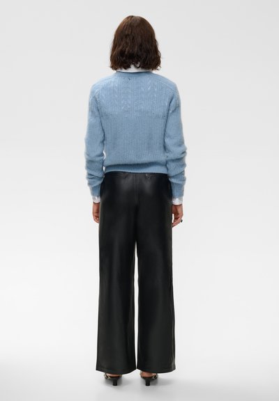 Femme debout de dos, portant un pull bleu clair, des poignets de chemise blancs, un pantalon noir à jambes larges et des chaussures noires à talons bas.