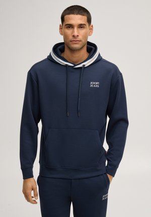 Mann trägt navyblauen Hoodie und passende Hose mit "JOOP! JEANS" Logo auf Brust und Oberschenkel, steht vor einfarbigem Hintergrund.