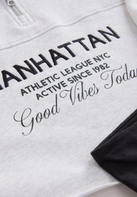 Grauer Pullover mit schwarzen Akzenten, versehen mit fettem und kursiv gedrucktem Text. Verfügt über einen halben Reißverschluss am Kragen und eine weiche Textur.