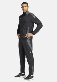 Schwarzer Sportanzug mit hellgrauen Akzenten, eingekleidete Vorderseite und Dreistreifen-Design an Ärmeln und Hosen. Getragen mit schwarzen Sneakers.