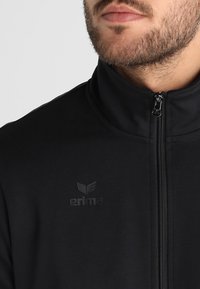 Veste zippée noire en tissu lisse, avec un col montant et un logo en relief discret sur la poitrine.