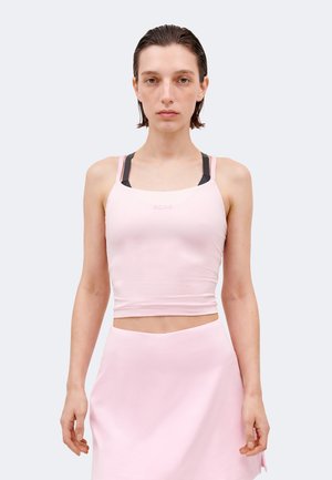 Kobieta z krótkimi, mokrymi włosami, ubrana w jasnoróżowy krótki top na czarnym sportowym biustonoszu i pasującą różową spódnicę.