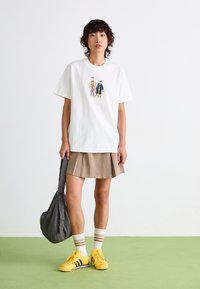 Hvit bomull T-skjorte med grafikk av tre tegneseriefigurer; kombinert med en beige folderkjole, gule sneakers og en gråmønstret veske.