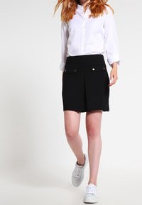 Falda negra con pliegues y bolsillos frontales, hecha de una tela suave, emparejada con una camisa blanca con botones que presenta patrones sutiles, y zapatillas blancas.