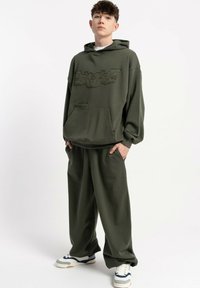 Młody mężczyzna stoi z rękami w kieszeniach, ubrany w oliwkowo-zielony duży hoodie oraz luźne odpowiadające spodnie, a na nogach ma białe sneakersy.