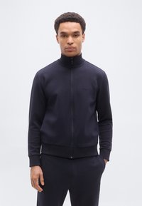 Άνδρας που φοράει μαύρο μπουφάν με φερμουάρ και ταιριαστά παντελόνια, στέκεται όρθιος με το ένα χέρι στην τσέπη, μπροστά σε ένα απλό φωτεινό φόντο.