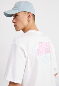 Balta marškinėliai su rožinės ir mėlynos spalvos grafika ant nugaros, kurioje užrašyta "ADIDAS ORIGINAL" ir "1972". Šviesiai mėlynas kepurė su lenkta kraštu.