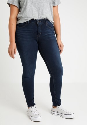 Person wearing dark blue skinny jeans, grey t-shirt, and white low-top sneakers, standing against a plain light background.