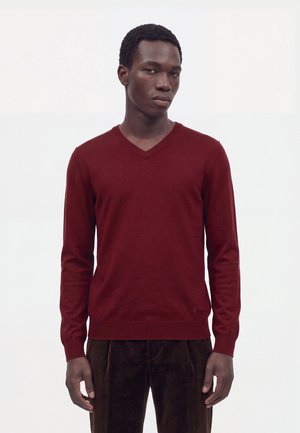 Un mannequin masculin se tient devant un fond blanc uni, vêtu d’un pull bordeaux à col en V et d’un pantalon foncé.