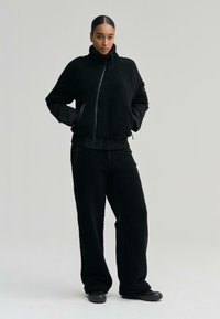 Černý fleecový svetr s vysokým límcem a zipem, spárovaný s širokými kalhotami ve stejném odstínu. Hladká textura, volný střih a minimalistické designové detaily.