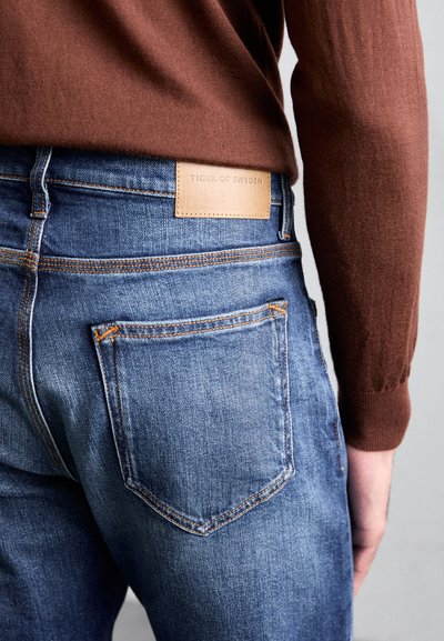 Jean's de ganga azul-escuro com uma etiqueta de couro que diz "TIGER OF SWEDEN." As características incluem costura em cobre e dois bolsos traseiros com um formato pontiagudo.