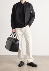 Veste bomber noire avec poches zippées, associée à un polo noir, un jean crème et un sac en cuir noir. Textures lisses et design structuré.
