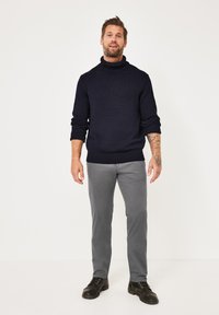 Navy gestrickter Rollkragenpullover mit gerippten Bündchen und Saum, kombiniert mit grauen Hosen und schwarzen Schnürschuhen, stehend vor einem schlichten Hintergrund.