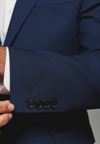 Morski niebieski męski żakiet z rękawem, wykonanym z teksturowanej tkaniny, z czterema czarnymi guzikami z detalami. Widoczny mankiet białej koszuli pod spodem.