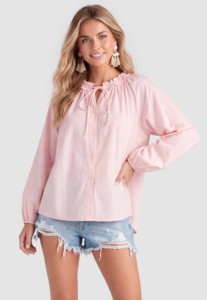 Frau trägt eine hellrosa Bluse mit langen Ärmeln und Rüschen am Ausschnitt sowie blaue, ausgewaschene Denim-Shorts, lächelnd mit Hand in der Hüfte.