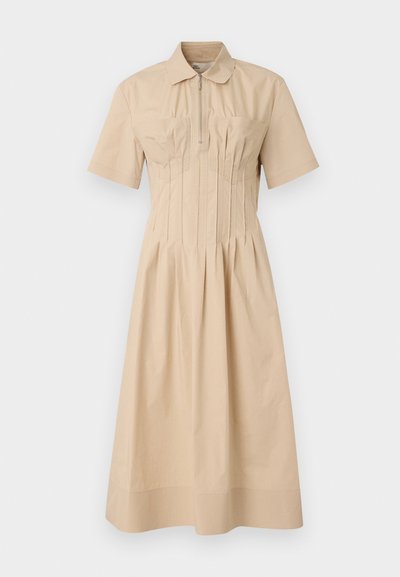 Beige lyhythihainen mekko, jossa on vetoketjukiinnitys, rypytetty yksityiskohta rinnan kohdalla ja liehuva hame. Valmistettu sileästä kankaasta.