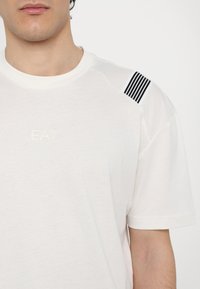 Balta T-kreklā ar apaļu kakla izgriezumu, kurā ir melna svītru detaļa uz pleca. "EA7" logo gaišā krāsā iespiests uz krūtīm.