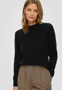 Schwarzer, gerippter Strickpullover mit langen Ärmeln und rundem Halsausschnitt, kombiniert mit braunen karrierten Hosen mit hoher Taille und Knopfdetails.
