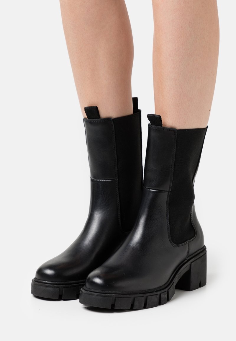 Bottes en cuir noir mi-mollet avec talons épais et languettes de tirage portées sur des jambes nues sur un fond clair uni.
