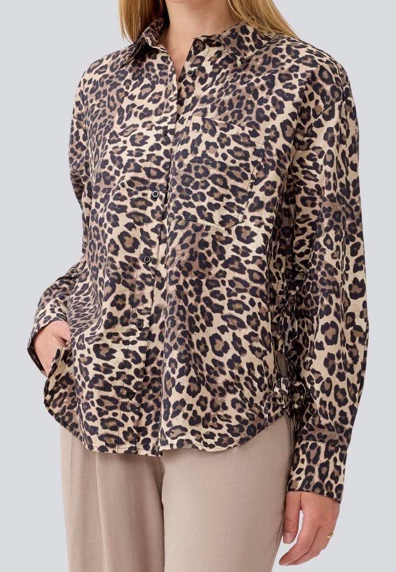 Femme portant une chemise à manches longues à boutons avec imprimé léopard et une poche avant, ainsi qu'un pantalon beige, la main reposant dans la poche.