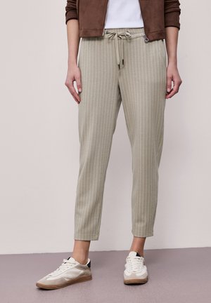 Osoba ubrana w beżowe spodnie w prążki ze sznurkiem, do tego białe trampki i brązowa kurtka, stojąca na szarej podłodze.