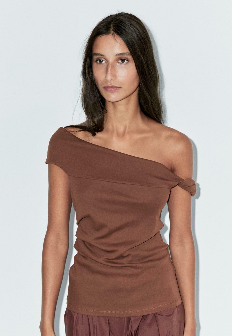 Top marrone, scollato sulle spalle, realizzato in tessuto morbido con dettaglio a nodo su una manica e texture drappeggiata lungo il corpo, design aderente.