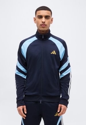 Uomo che indossa una giacca sportiva con cerniera blu navy e azzurro chiaro e pantaloni coordinati con logo dorato, in piedi davanti a uno sfondo chiaro e uniforme.