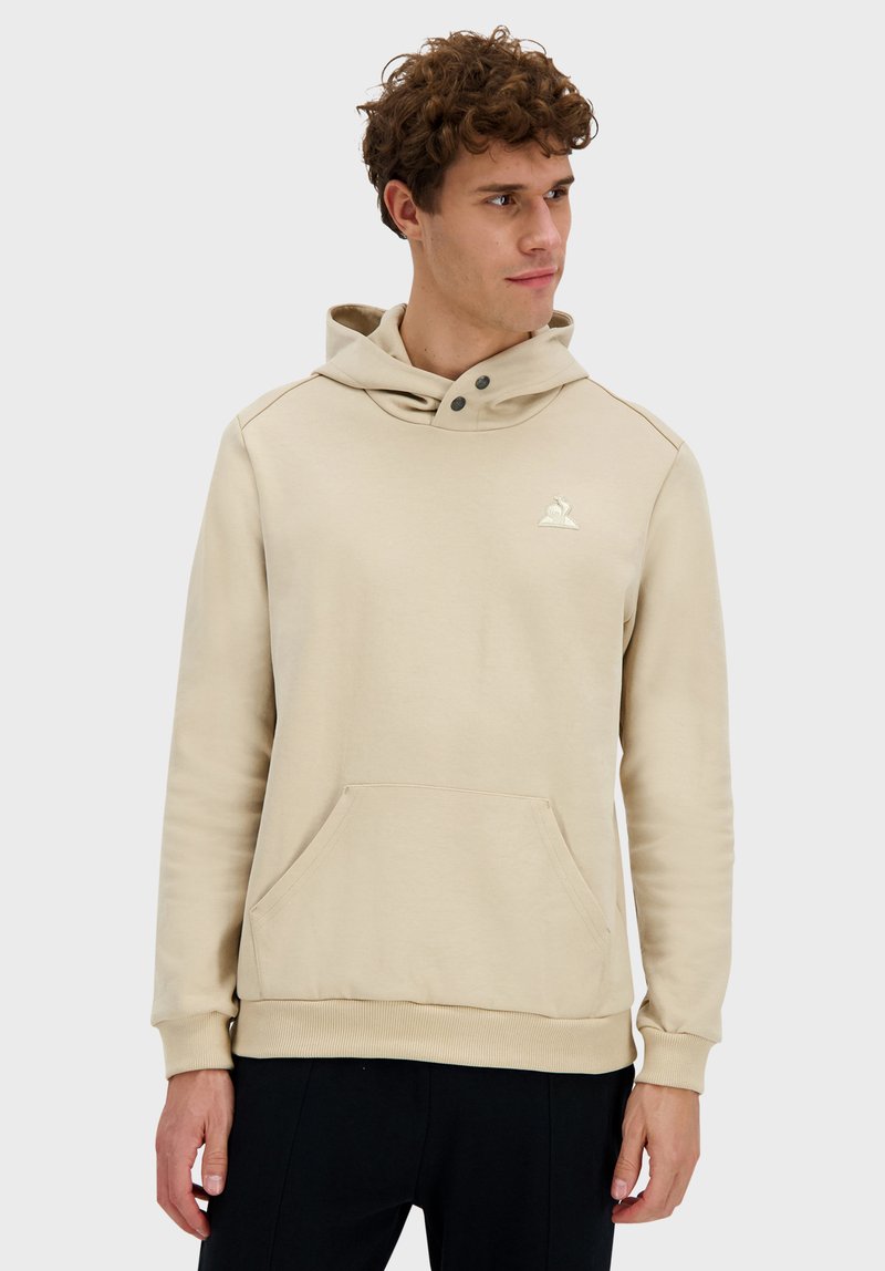 Sudadera con capucha beige de tela suave, con un bolsillo tipo canguro, detalles de botones falsos en el cuello y un pequeño logo en el pecho.