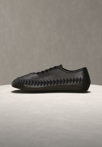 Chaussure en cuir noir avec coutures décoratives noires le long des côtés et du dessus, présentée sur une surface gris clair sur fond gris uni.