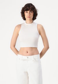Bílý žebrovaný crop top s imitovaným límečkem, v kombinaci s bílými kalhotami s vysokým pasem a předním knoflíkem a detailem na zavázání.