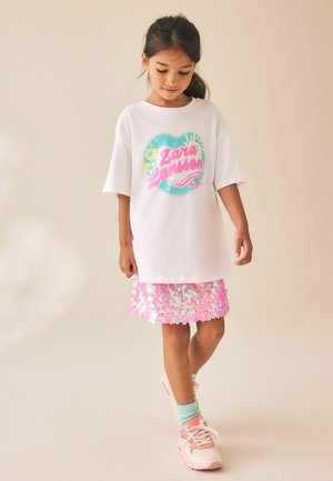 Jong meisje kijkt naar beneden, draagt een oversized wit Zara Larsson t-shirt, een roze pailletten rok, mintgroene sokken en roze sneakers, staat op een beige vloer.