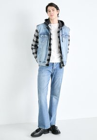 Denimväst med två framfickor över en svart- och vitrutig skjorta, kombinerad med ljusblå jeans och svarta skor.