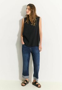 Zwart mouwloos topje, losse fit; blauwe denim jeans opgerold bij de boorden; zwarte slip-on sandalen met gouden accenten. Glad materiaal en casual ontwerp.